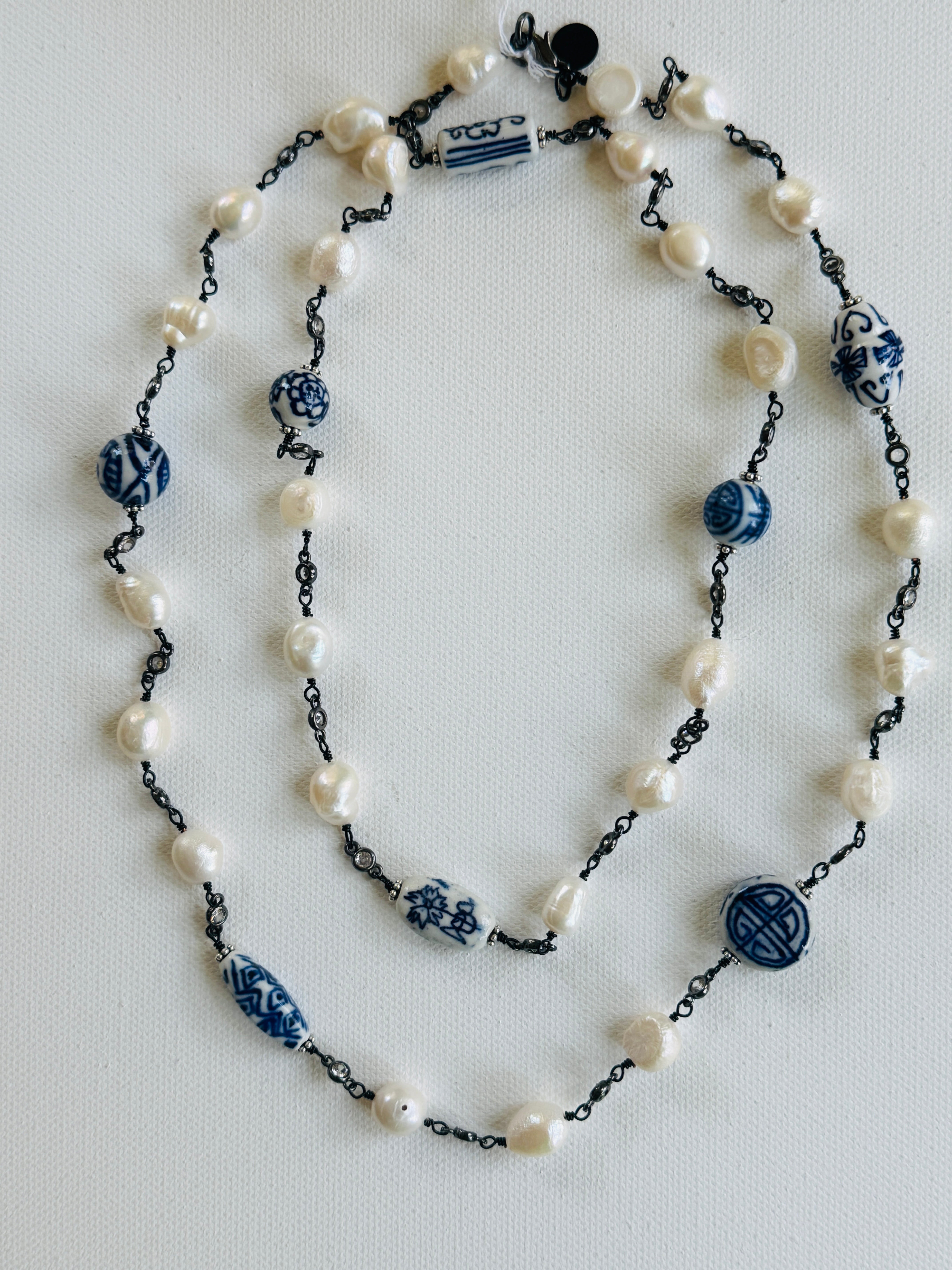 A double-strand beaded necklace featuring white freshwater pearls, gold-tone chain links, and blue-and-white ceramic beads with traditional geometric patterns, laid out on a light gray background.