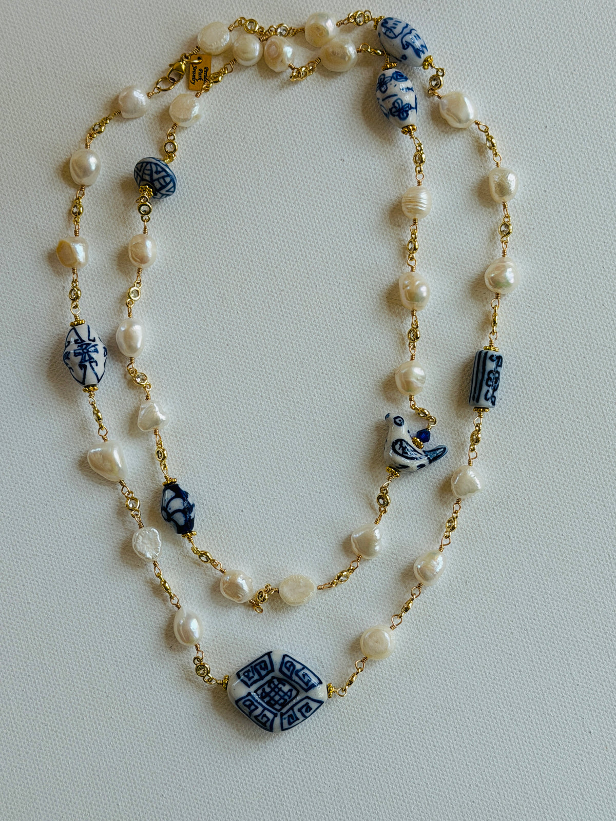 A double-strand beaded necklace featuring white freshwater pearls, gold-tone chain links, and blue-and-white ceramic beads with traditional geometric patterns, laid out on a light gray background.