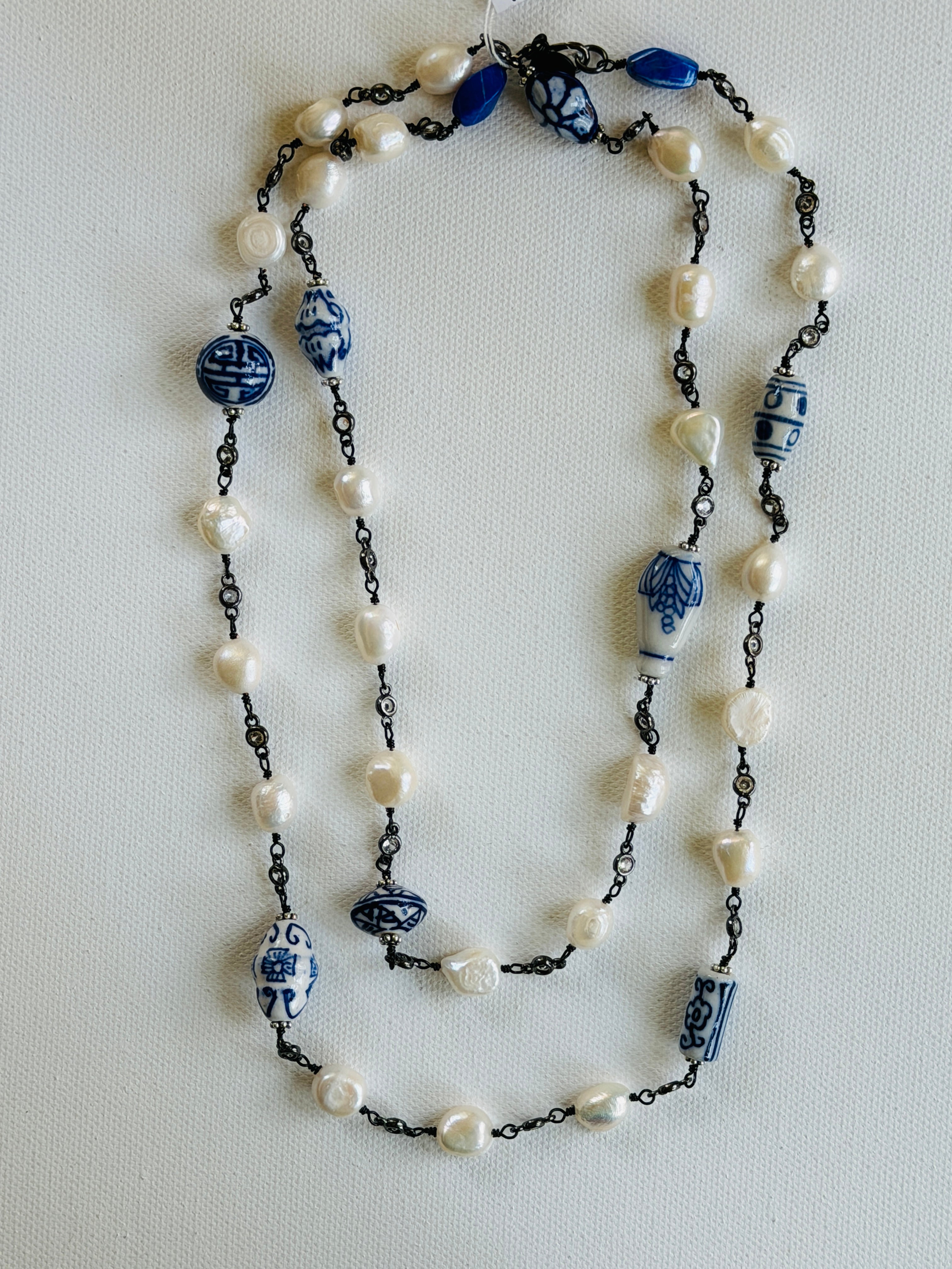 A long beaded necklace featuring white freshwater pearls, blue and white ceramic beads, and dark metal chain links, laid flat on a light textured background.