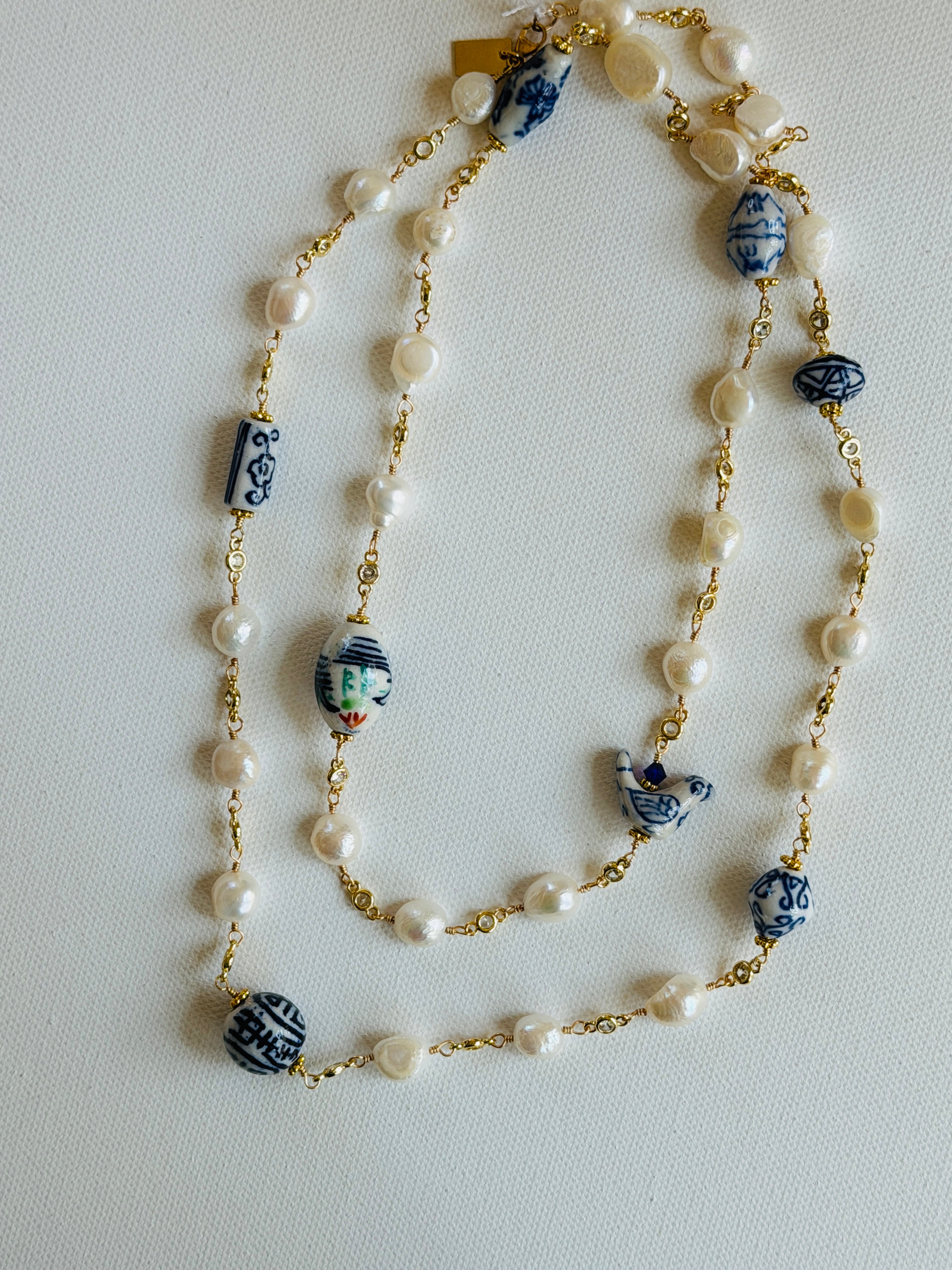 A long beaded necklace featuring white faux pearls, gold-tone chain links, and blue-and-white ceramic decorative beads with hand-painted details, laid out on a light off-white background.