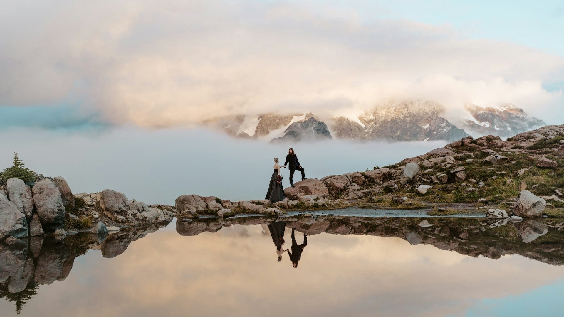 Two people standing on a rocky outcrop with a mountainous landscape reflected in a calm body of water.