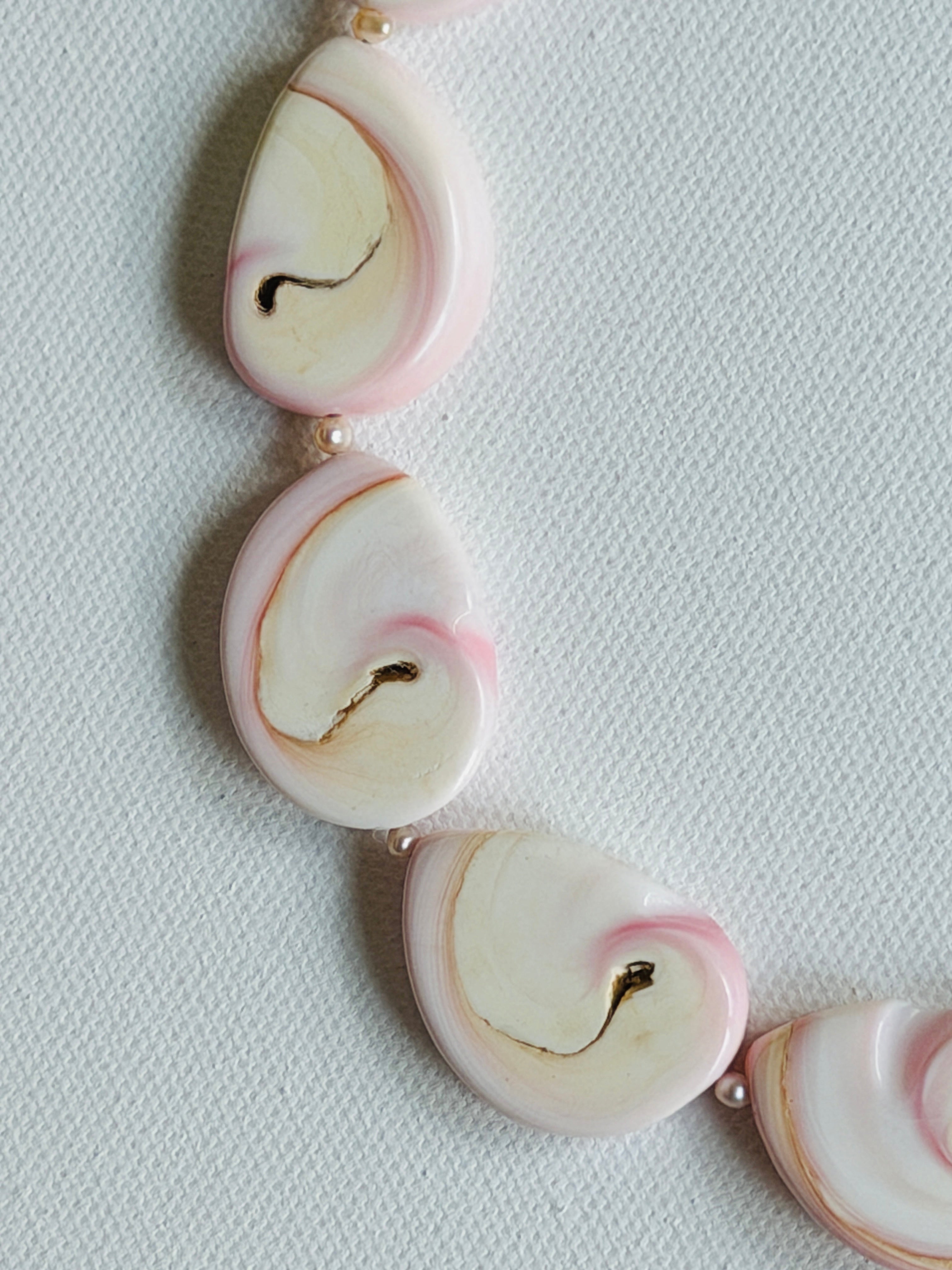 A close-up of a necklace featuring organic-shaped Conch Shell slices in soft pink, separated by small pink pearl spacer beads on a textured white background.