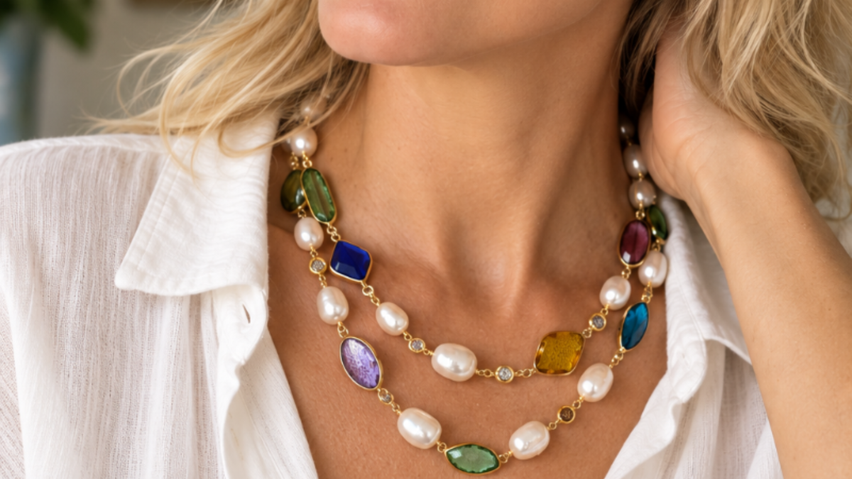 A close-up lifestyle shot of a woman wearing a multi-strand statement necklace with white pearls and colorful faceted crystal accents, paired with a white button-down shirt.