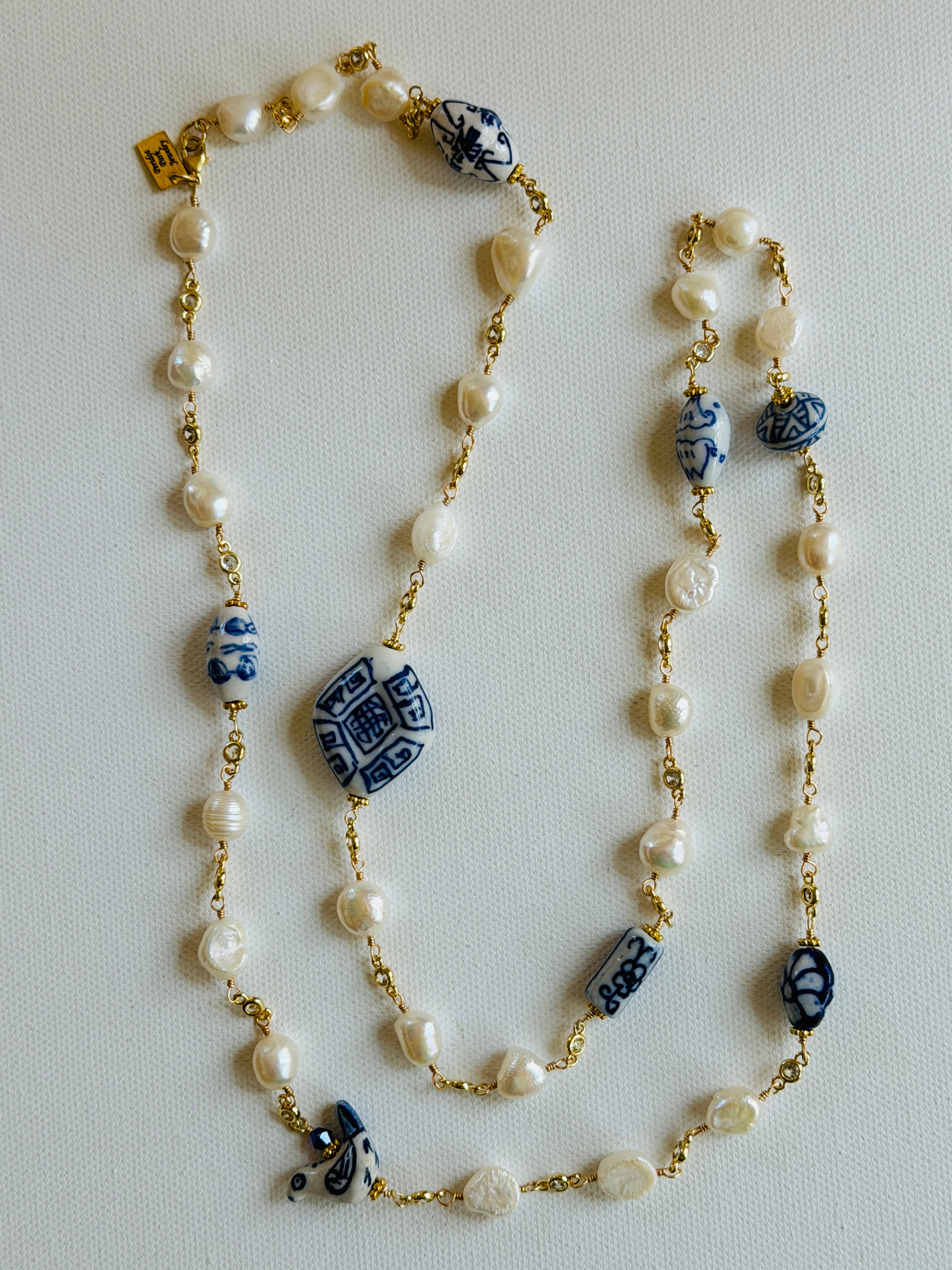 A long beaded necklace laid flat on a light background, featuring white faux pearls, gold-tone chain links, and blue-and-white patterned ceramic beads.