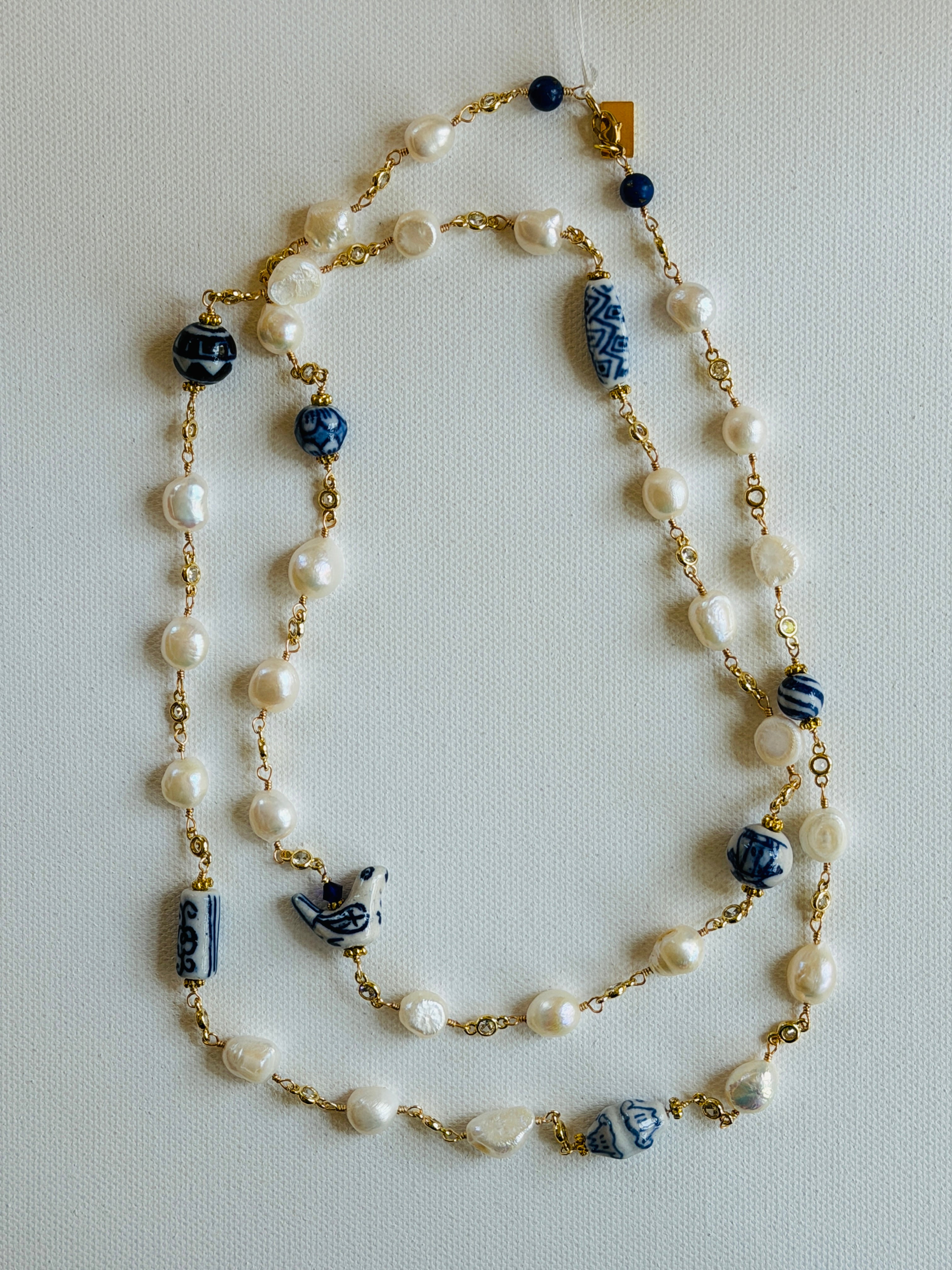 A long beaded necklace featuring white freshwater pearls, blue and white patterned ceramic beads, and gold-tone chain links, displayed on a light gray textured background.