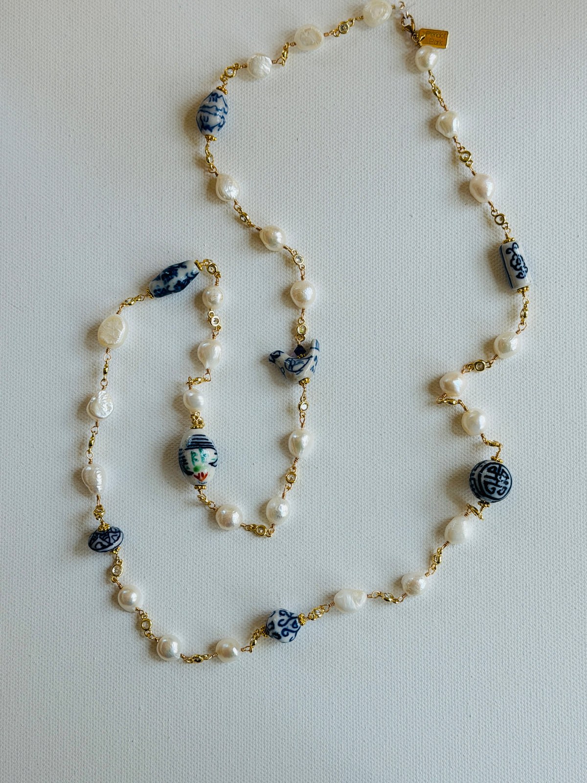 A long beaded necklace with white faux pearls, blue and white ceramic patterned beads, and gold-tone chain links, laid out on a light gray background.