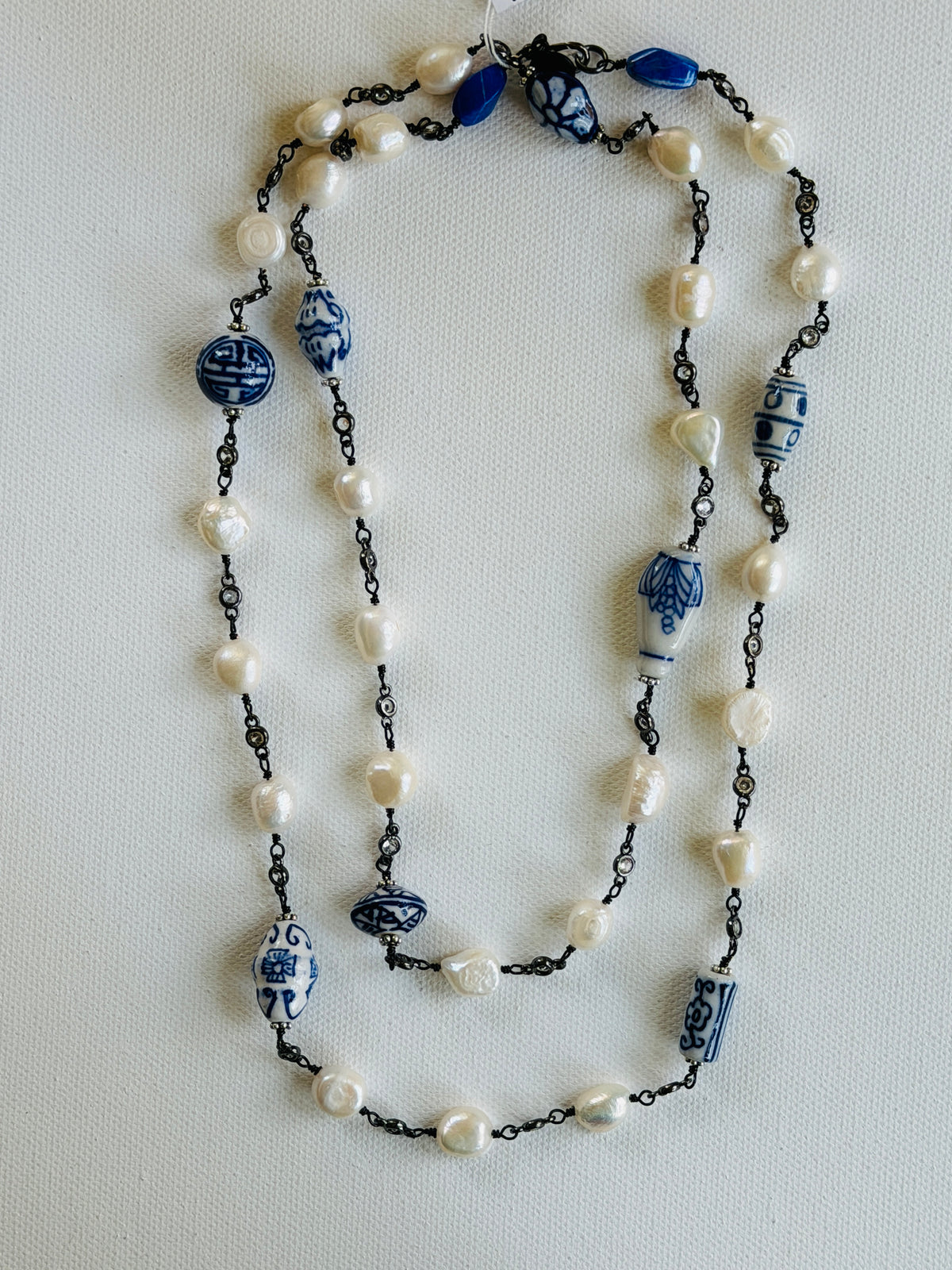 A long beaded necklace featuring white freshwater pearls, blue and white ceramic beads, and dark metal chain links, laid flat on a light textured background.